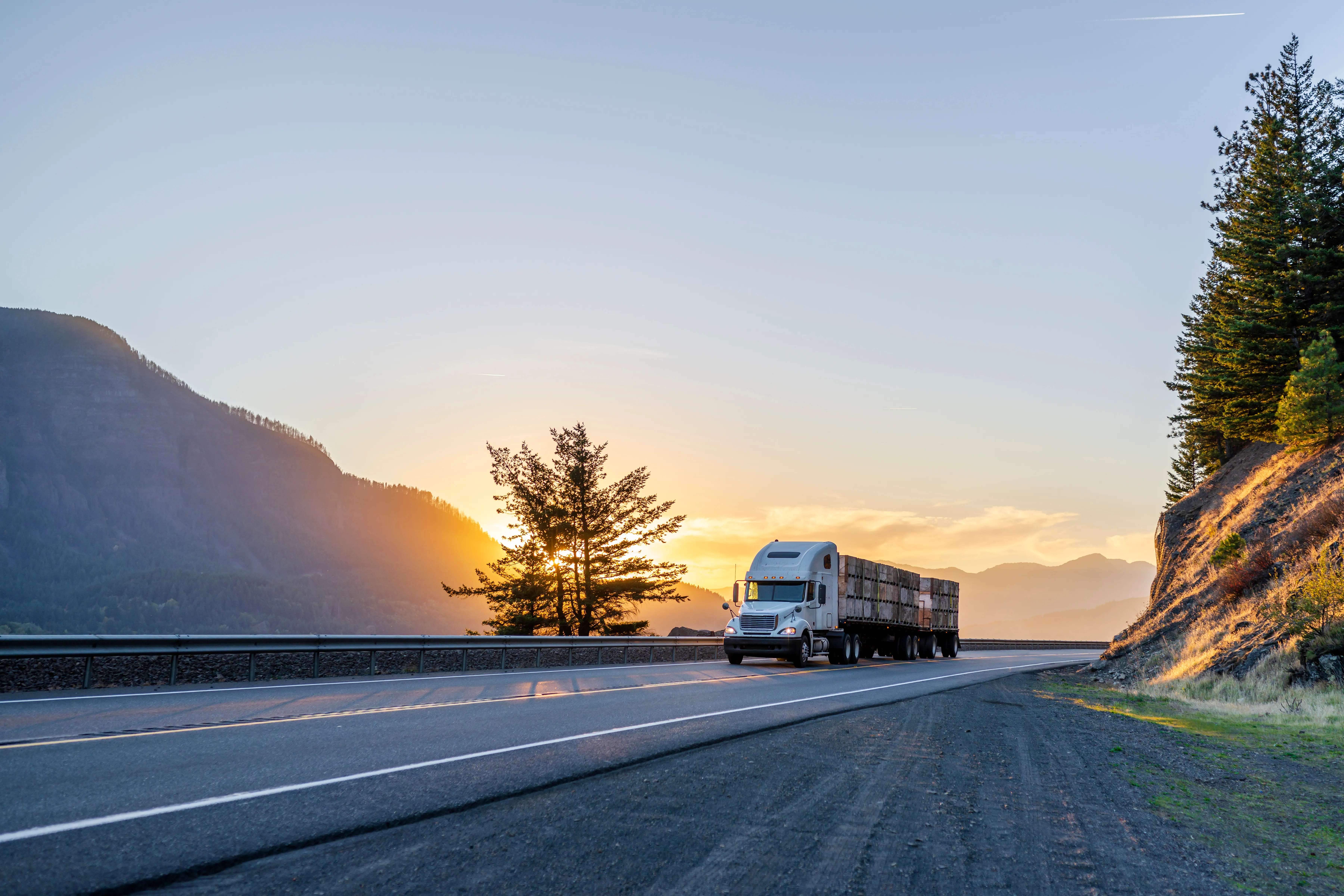 Semi truck hauling a loaded flatbed trailer through mountains at sunset