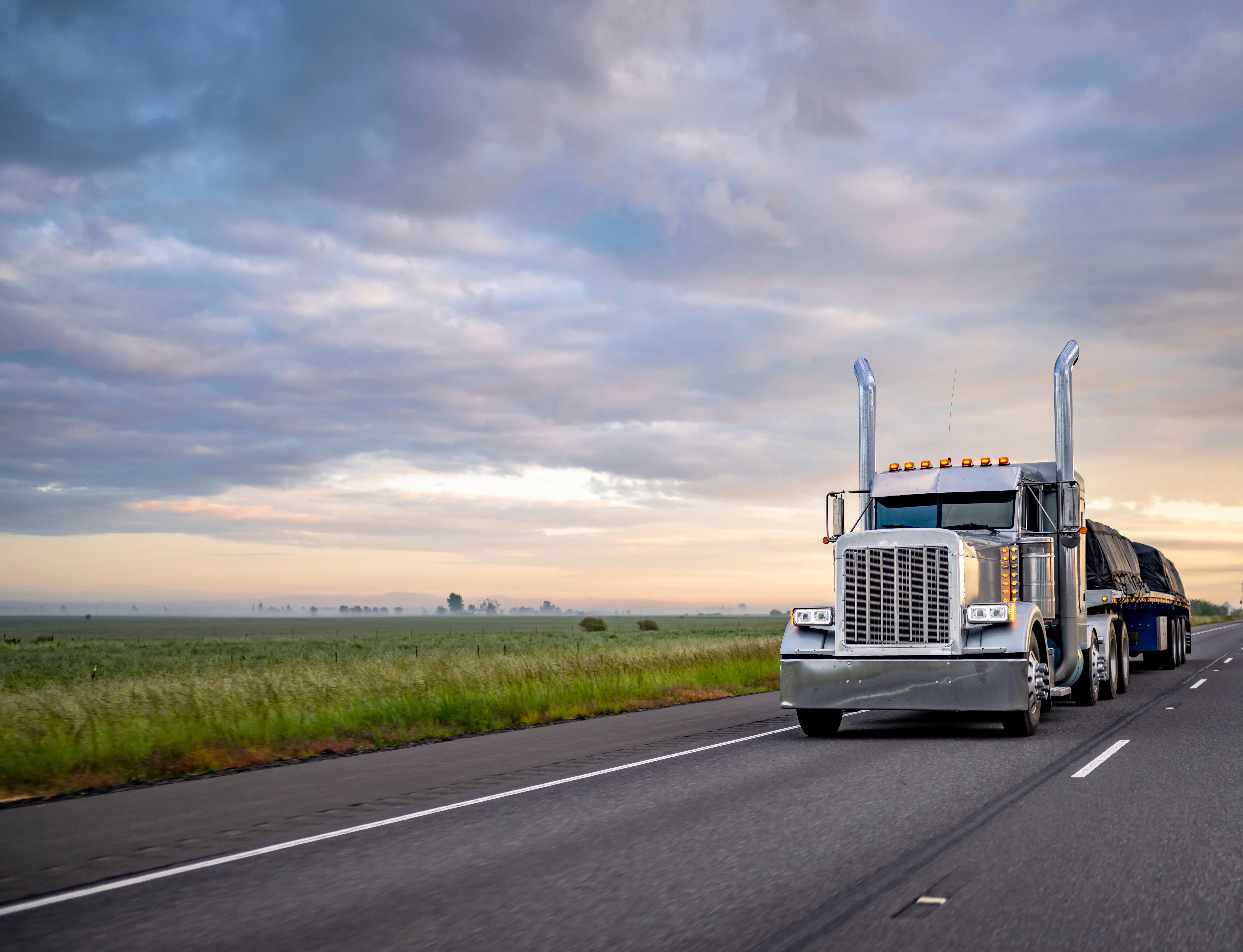 Silver semi truck hauling a tarped flatbed load down the highway at dawn